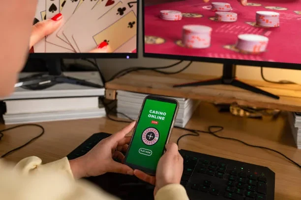 একটি মোবাইল ফোনে ‘CASINO ONLINE’ লেখা অনলাইন ক্যাসিনো স্ক্রিন দেখা যাচ্ছে; পেছনের দুটি মনিটরে কার্ড ও রুলেটের দৃশ্য রয়েছে।
